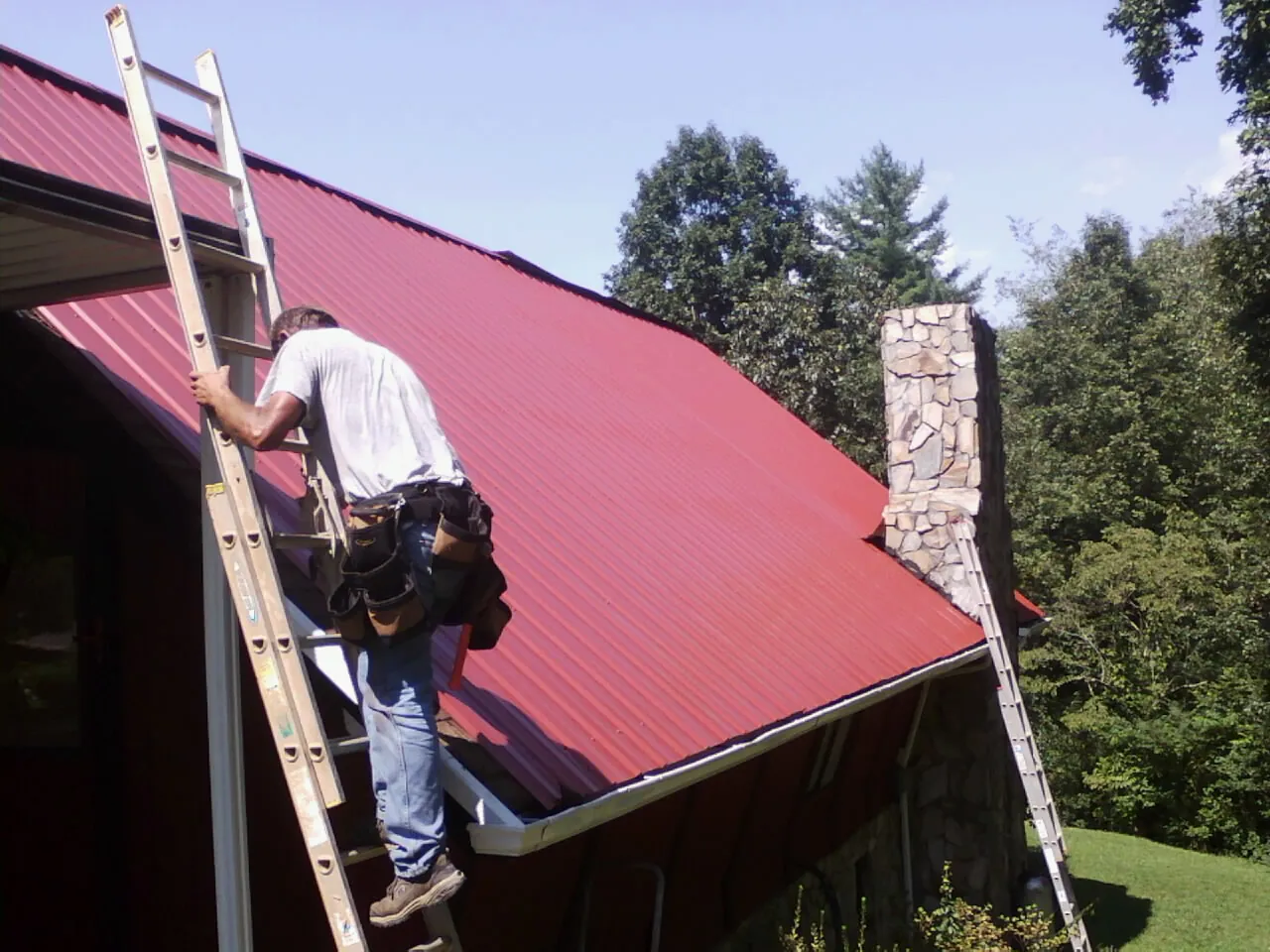 Roofer on a ladder beside a red metal roof and stone chimney — Soffit & Fascia Repair by Roofing Brothers in Haddonfield